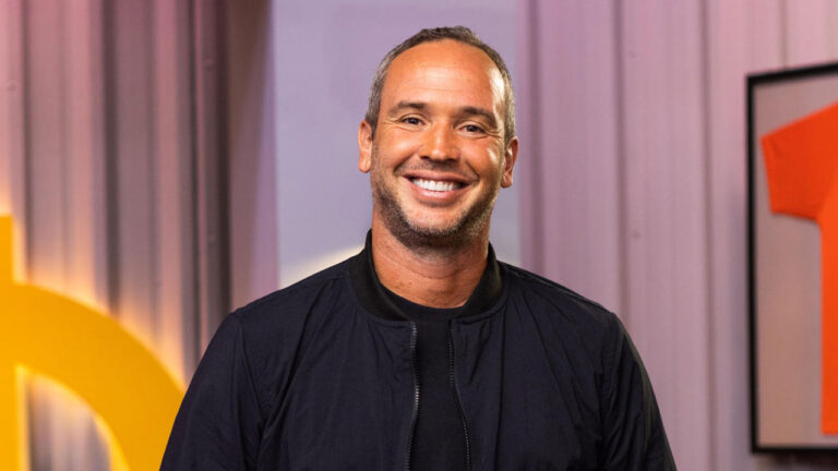 Caio Ribeiro com jaqueta preta e camiseta escura sorrindo de forma aberta para a câmera em estúdio da Globo