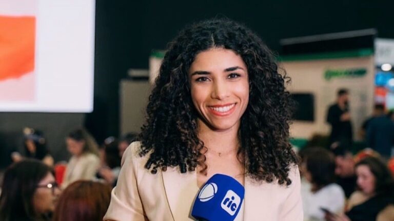 Repórter jovem com cabelos cacheados escuros sorri enquanto segura um microfone azul de uma emissora. Ela veste blazer claro e está em um ambiente interno movimentado, com várias pessoas desfocadas ao fundo