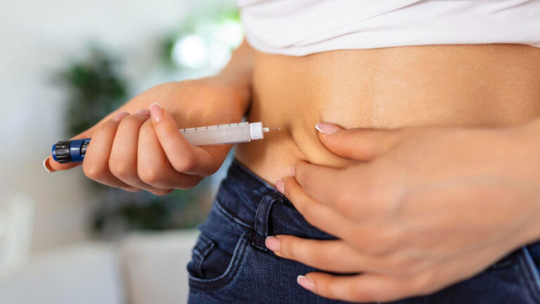 Mulher aplica injeção na região abdominal utilizando uma caneta emagrecedora, segurando uma dobra de pele com a outra mão