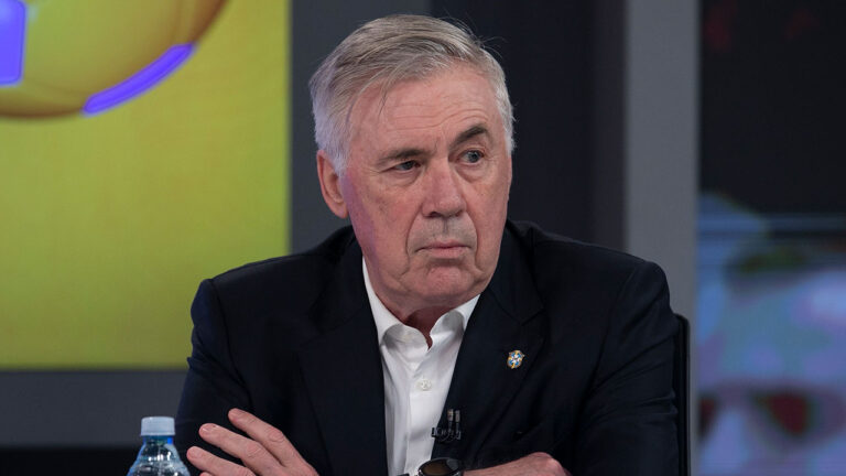 Técnico Carlo Ancelotti de camisa social branca e terno preto com olhar sério e braços cruzados durante entrevista para Record