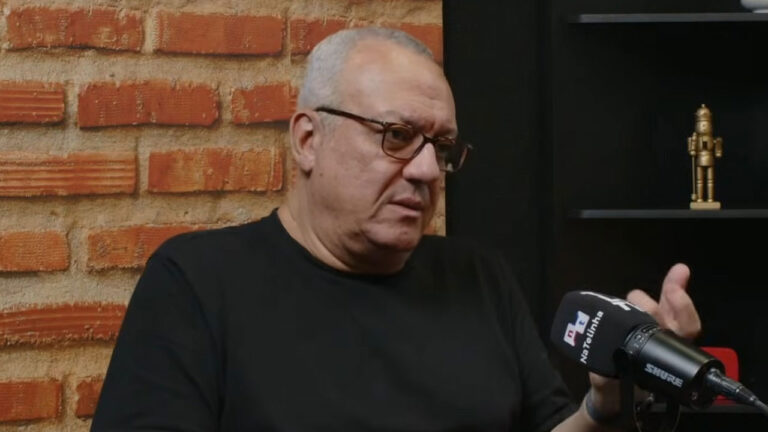 Carlos Henrique Nascimento, de óculos e camisa preta, gesticula enquanto fala ao microfone em entrevista