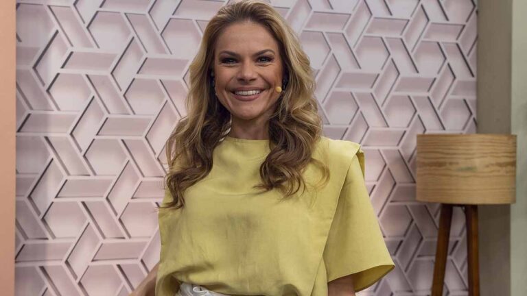 Uma mulher aparece em um estúdio bem iluminado, sorrindo diretamente para a câmera. Ela tem cabelos loiros, longos e ondulados, usa uma blusa amarela de mangas curtas e um microfone de lapela preso próximo ao rosto. O fundo é uma parede decorativa com padrão geométrico em tons claros, e ao lado há um abajur com base de madeira e cúpula clara, reforçando o ambiente de estúdio ou programa de TV com clima acolhedor e profissional.