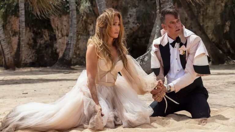 Homem e mulher ajoelhados na areia de uma praia, vestidos com trajes de casamento. A mulher usa um vestido claro e volumoso, com expressão tensa, enquanto o homem, de smoking parcialmente desalinhado, segura a mão dela e olha para o lado com preocupação. Ao fundo, há palmeiras e formações rochosas.