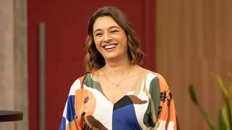 Mulher sorridente com cabelo castanho na altura dos ombros usa vestido colorido com estampas geométricas em tons de laranja, verde, azul e branco, e microfone de lapela preso ao rosto, em ambiente interno com fundo vermelho e madeira clara