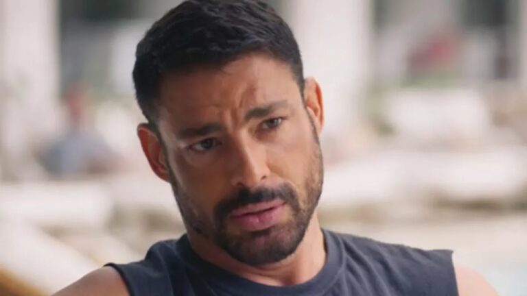 Homem adulto em primeiro plano com barba curta e cabelo escuro usando camiseta sem mangas escura com expressão séria e olhar levemente inclinado para baixo em ambiente externo desfocado com colunas claras e luz natural sugerindo área aberta ou varanda