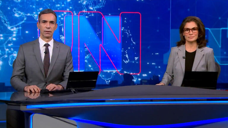César Tralli e Renata Vasconcellos de óculos apresentam o Jornal Nacional na bancada com cenário azul e mapa mundi ao fundo