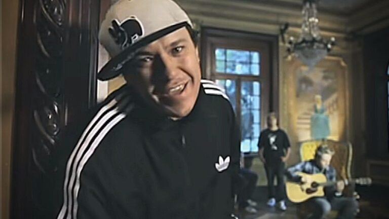 Homem usando boné e agasalho esportivo preto com listras brancas aparece em primeiro plano dentro de um salão antigo iluminado, olhando para a câmera com expressão animada. Ao fundo, outras pessoas estão sentadas e tocando violão, diante de uma grande janela e de um quadro decorativo, criando clima de ensaio musical.