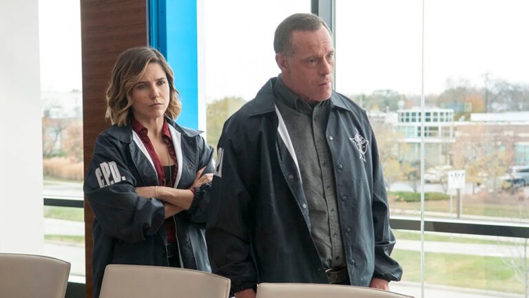 Cena da série Chicago P.D., exibida pela Record, mostra dois detetives usando jaquetas escuras com o emblema da polícia de Chicago (C.P.D.) em uma sala iluminada por janelas amplas. A mulher está de braços cruzados, com expressão séria, enquanto o homem observa atentamente à frente, sugerindo uma discussão ou investigação em andamento