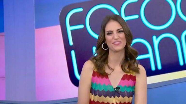 Uma mulher sorri para a câmera em um estúdio de televisão. Ela tem cabelos castanhos ondulados na altura dos ombros, usa brincos de argola e um vestido sem mangas com estampa colorida em tons de rosa, verde, amarelo e roxo. Ao fundo, há um painel iluminado com letras grandes em azul e rosa, sugerindo o nome de um programa de TV.