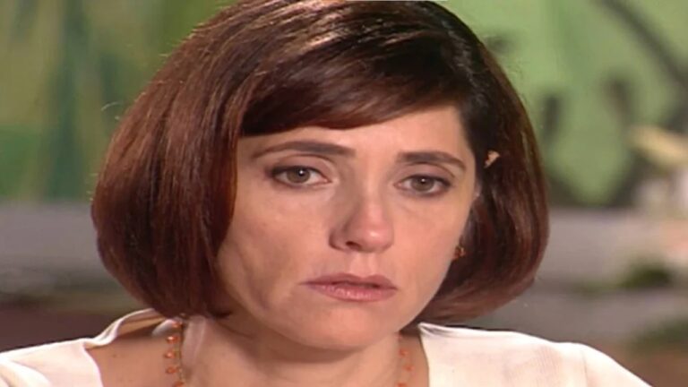 Mulher de cabelo castanho curto, com expressão abatida e olhar entristecido, aparece em close. Ela usa blusa clara e um colar delicado, com fundo desfocado em tons suaves.