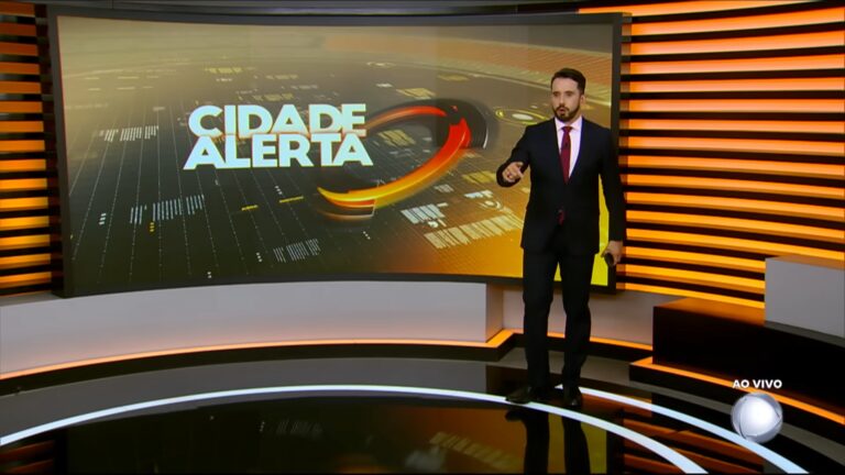 A imagem mostra Dionísio Freitas no estúdio do programa Cidade Alerta, da Record. Ele está de terno escuro e gravata vermelha, segurando um microfone, em frente a um grande telão que exibe o nome do telejornal. O cenário tem iluminação em tons de laranja e amarelo, com o logotipo da emissora visível no canto inferior direito e a indicação de transmissão ao vivo