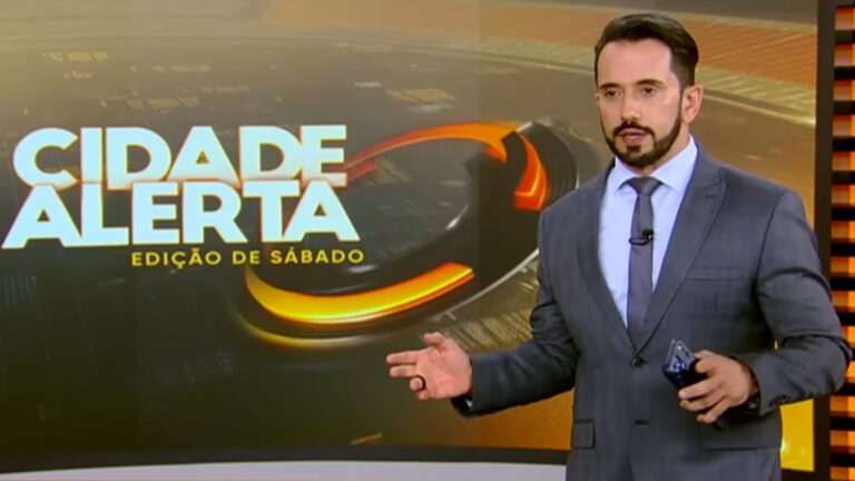 A imagem mostra Dionísio Freitas no estúdio do Cidade Alerta – Edição de Sábado. Ele veste um terno cinza com camisa clara e gravata, segurando um celular em uma das mãos. O cenário tem tons alaranjados e amarelos, com o logotipo do programa exibido em um grande painel ao fundo