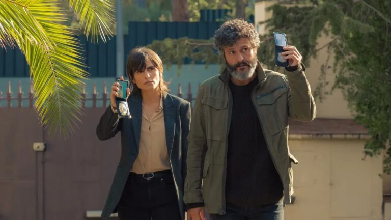 Duas pessoas caminham ao ar livre em frente a um portão residencial. À esquerda, uma mulher de cabelo curto e franja, vestindo blazer escuro e camisa clara, segura um celular aberto mostrando um distintivo. À direita, um homem de barba e cabelo grisalho cacheado, usando jaqueta verde, também levanta um celular aberto com identificação. Ambos têm expressão séria, como se estivessem se apresentando oficialmente. Ao fundo, há árvores, um muro e vegetação, com uma palmeira em primeiro plano.