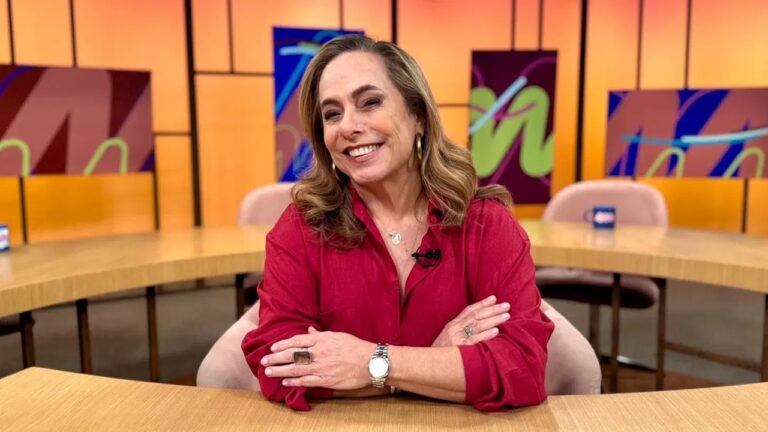 Apresentadora sorridente de blusa vermelha posa apoiada em mesa de estúdio colorido do programa Sem Censura