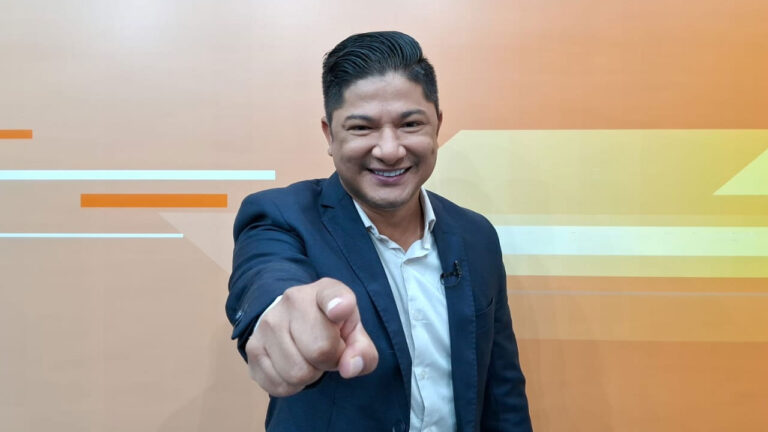 Cleytton Rasec sorri e aponta o dedo para a câmera usando terno azul em cenário laranja de estúdio de televisão