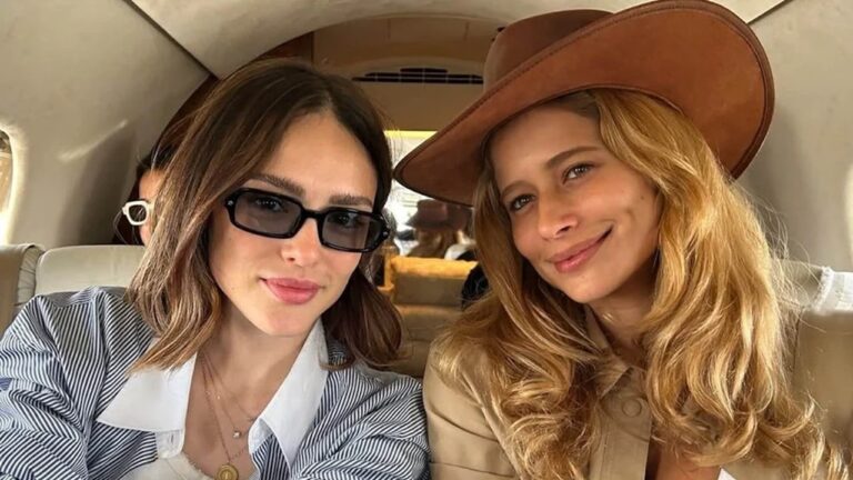 Duas mulheres sentadas lado a lado dentro de um avião pequeno, sorrindo para a câmera. A da esquerda usa óculos escuros retangulares, camisa listrada azul e branca e colares dourados, enquanto a da direita tem cabelos loiros soltos, usa chapéu marrom de aba larga e blusa bege clara.