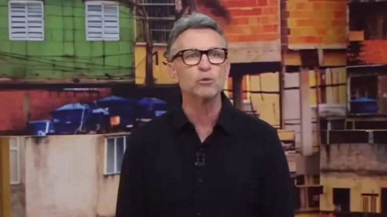 Homem de cabelos grisalhos e óculos de aro grosso, vestindo camisa preta, aparece em frente a um painel com imagem de construções urbanas coloridas, como casas e prédios simples. Ele olha para a câmera com expressão séria