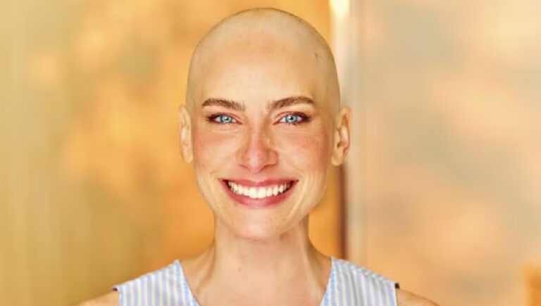 Pessoa adulta em close frontal com cabeça raspada pele clara olhos azuis sorriso aberto mostrando os dentes sobrancelhas finas maquiagem leve usando blusa sem mangas clara com listras finas em tons suaves fundo desfocado em tons quentes de bege e dourado com iluminação suave que destaca o rosto e transmite sensação de serenidade e acolhimento