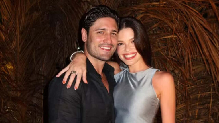 Casal posando abraçado em ambiente externo à noite. Eles sorriem para a câmera; o homem veste camisa preta e a mulher usa vestido prateado sem mangas. O fundo é composto por folhagens secas iluminadas artificialmente, destacando o clima festivo da cena