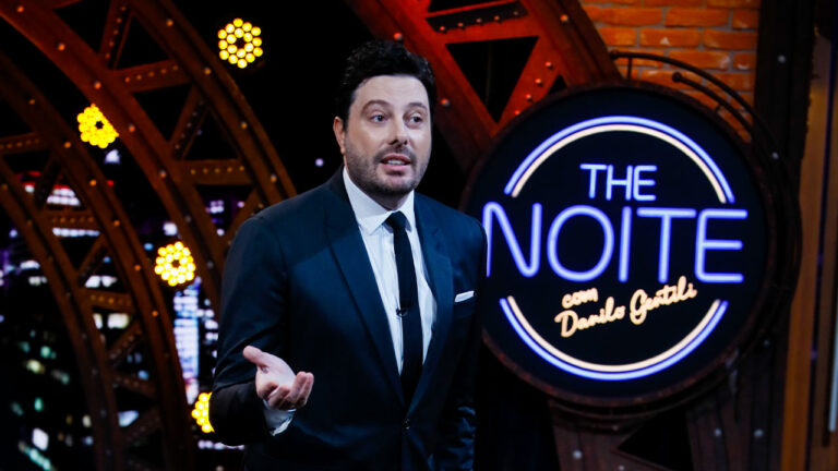 Danilo Gentili de terno escuro gesticula com a mão aberta à frente do logotipo luminoso do programa The Noite nos estúdios do SBT