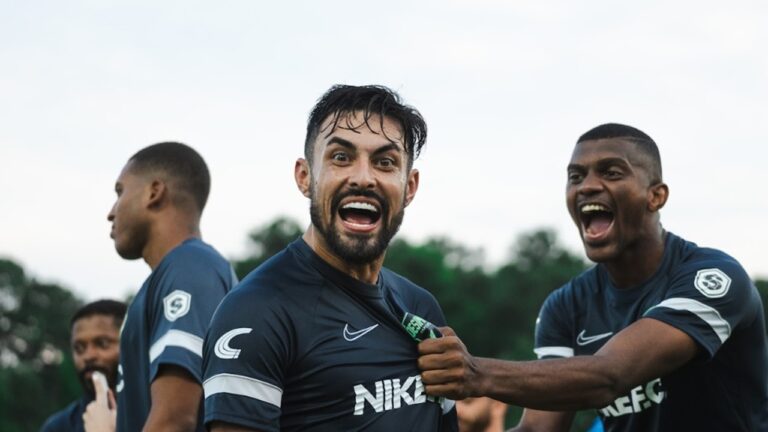 Jogadores de futebol comemoram em campo com uniforme escuro da Nike FC com atleta ao centro puxando a camisa e companheiros gritando ao redor