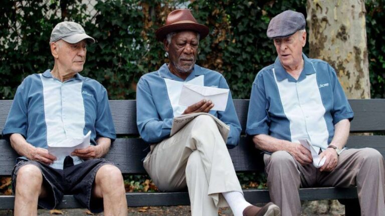 Três homens idosos sentam em um banco de parque vestidos com camisas azul-claro semelhantes, segurando papéis e conversando enquanto árvores e folhagem aparecem ao fundo