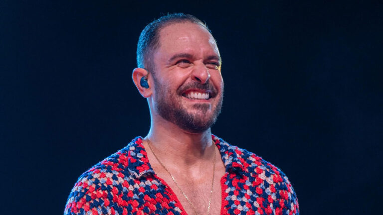 Diogo Nogueira sorri com os olhos fechados usando camisa de crochê colorida e fone de retorno durante apresentação em show