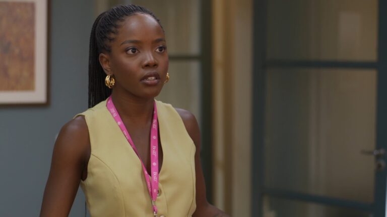 A imagem mostra a personagem Leona, interpretada por Clara Moneke, em Dona de Mim. Ela é uma mulher em um ambiente interno, aparentando estar em um escritório. Ela veste um colete bege e usa um crachá rosa pendurado no pescoço, além de brincos dourados. A expressão em seu rosto é séria e atenta, com o fundo desfocado revelando portas de vidro e uma parede azul acinzentada