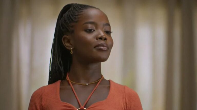 Mulher com expressão confiante e olhar altivo veste blusa laranja e colares finos, com o cabelo preso em tranças longas e o ambiente ao fundo desfocado por cortinas claras.