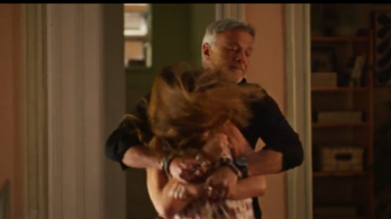 Homem de meia-idade abraça com força uma mulher em um corredor de casa, segurando-a pelos braços enquanto ela tenta se soltar; seus cabelos estão em movimento, sugerindo agitação. A cena ocorre em ambiente doméstico, com iluminação quente e estantes ao fundo, transmitindo tensão e conflito