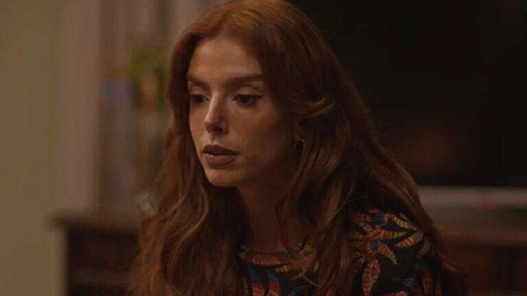 Mulher jovem com cabelos ruivos soltos aparece sentada em ambiente interno pouco iluminado, olhando para baixo com expressão séria e pensativa. Ela veste uma blusa estampada em tons quentes, e o fundo desfocado sugere um cenário doméstico.