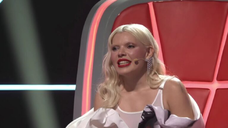 Mulher sentada em uma grande cadeira de encosto vermelho em um estúdio iluminado, usando microfone de testa, brincos brilhantes e batom vermelho. Ela sorri enquanto fala, vestindo roupa clara com tecido drapeado sobre os ombros. Linhas de luz no fundo sugerem um cenário de programa de TV