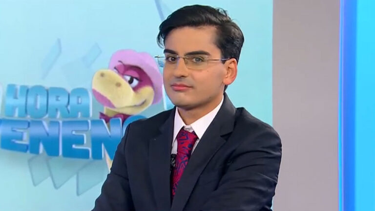 Dudu Camargo de terno, óculos e gravata vermelha no cenário do quadro A Hora da Venenosa com a cobra Judite ao fundo na Record