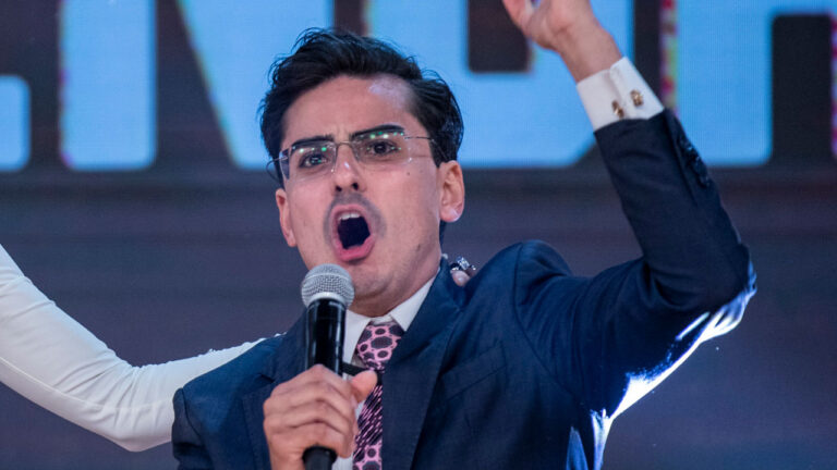 Dudu Camargo de terno e gravata rosa fala ao microfone com o braço erguido durante a final de A Fazenda 17