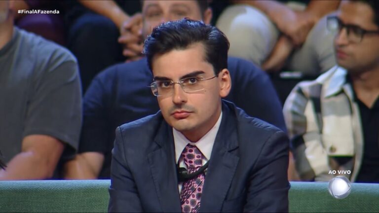 A imagem mostra Dudu Camargo, vestido de terno escuro, camisa branca e gravata rosa estampada, usando óculos e com expressão séria. Ele está sentado em um estúdio de TV, com outras pessoas desfocadas ao fundo, durante uma transmissão ao vivo identificada pelo logotipo da Record na final de A Fazenda 17