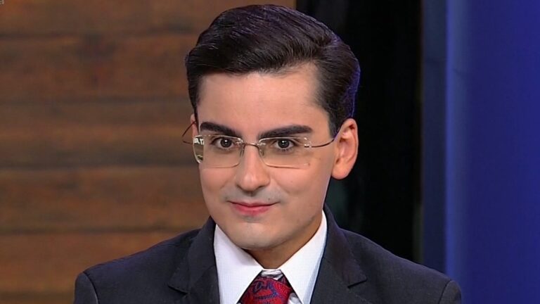 A imagem mostra Dudu Camargo, um homem jovem, de cabelos escuros penteados para trás, usando terno escuro, camisa branca e gravata vermelha estampada. Ele também usa óculos de aro fino e aparece sorrindo levemente em um estúdio de televisão, do programa Hoje em Dia, da Record