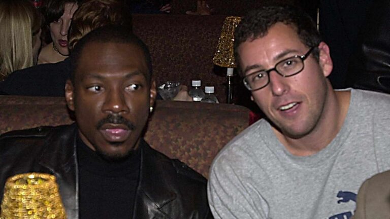 Dois homens sentados lado a lado em um ambiente interno com pouca luz, semelhante a um bar ou lounge. O homem à esquerda usa jaqueta de couro escura e camiseta preta, olhando para o lado. O homem à direita usa óculos e camiseta cinza, olhando para a câmera. Ao fundo, há mesas com luminárias douradas e outras pessoas conversando