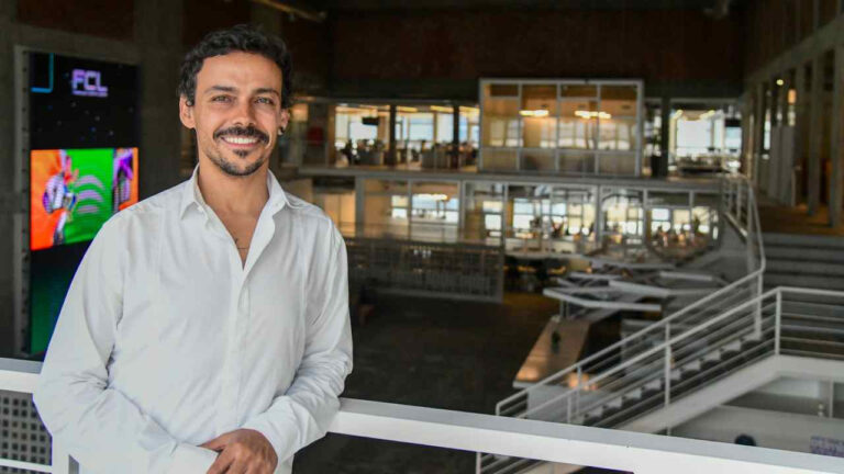 Homem sorrindo, de camisa branca, encostado em um corrimão dentro de um amplo espaço corporativo moderno. Ao fundo, há ambientes envidraçados, mesas de trabalho, iluminação interna e uma grande tela colorida exibindo imagens digitais.