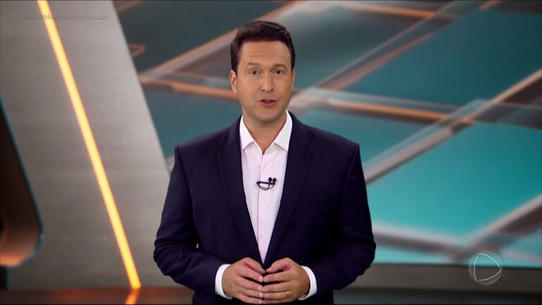 A imagem mostra Eduardo Ribeiro no estúdio do programa Domingo Espetacular, da Record. Ele está de terno escuro e camisa branca, com um microfone preso à lapela. No canto superior esquerdo da tela aparece a hashtag #DomingoEspetacular, e no canto inferior direito, o logotipo da emissora. O fundo tem um design geométrico moderno em tons de azul e laranja