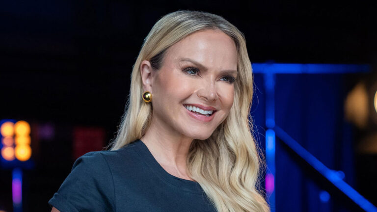 Eliana sorri de camiseta preta e cabelos loiros soltos em estúdio com iluminação azul e laranja desfocada ao fundo