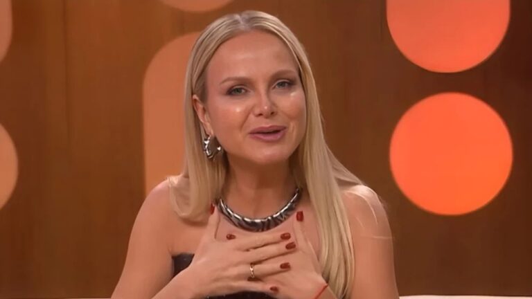 Apresentadora loira fala emocionada em estúdio de TV com mão no peito usando colar prateado e fundo com iluminação circular alaranjada