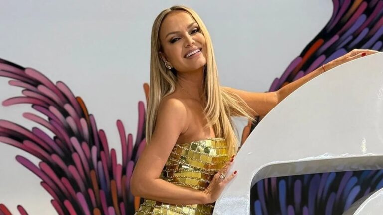 Imagem de Eliana, uma mulher sorridente em um cenário colorido; ela usa um vestido dourado brilhante e posa apoiada em uma estrutura branca, diante de um fundo com ilustrações em tons de roxo, rosa e laranja que lembram penas estilizadas.