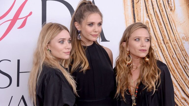 Três mulheres posam juntas em um evento, todas vestindo roupas pretas elegantes. A mulher ao centro está com o cabelo preso em um rabo alto; as duas nas laterais usam o cabelo solto e ondulado, cada uma com acessórios chamativos, como brincos grandes. Elas estão diante de um painel claro com letras grandes ao fundo