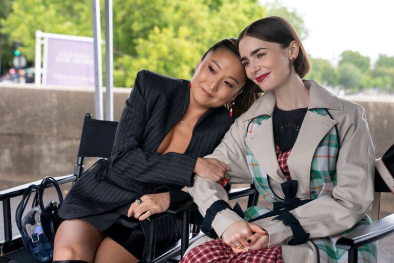 Emily Cooper e Mindy Chen sentadas em cadeiras de diretor apoiam a cabeça uma na outra em bastidores ao ar livre da série Emily in Paris