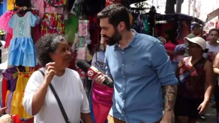 Repórter segura um microfone e entrevista uma mulher em um mercado popular ao ar livre. Eles estão cercados por barracas com roupas coloridas penduradas, enquanto outras pessoas circulam pelo local, caracterizando um ambiente movimentado de comércio de rua.