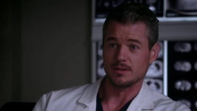 Homem de cabelo curto e barba rala usa jaleco branco e camiseta escura embaixo, aparecendo em ambiente hospitalar com exames de imagem pendurados em painel luminoso ao fundo