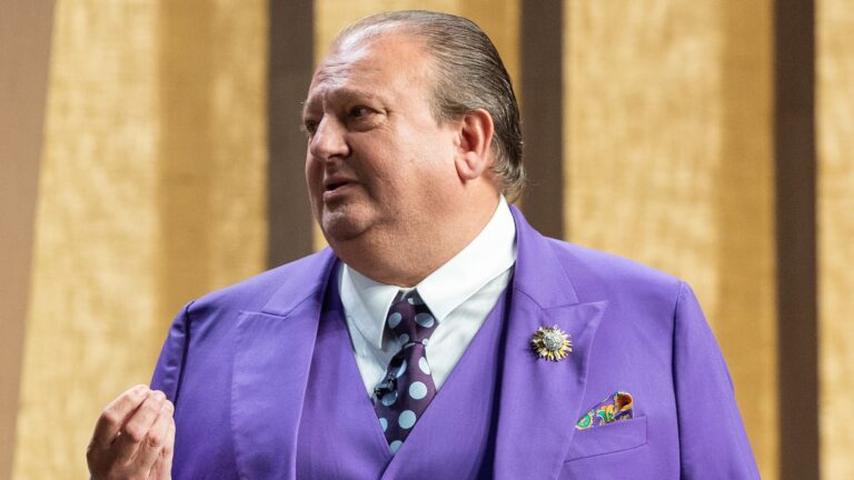 A imagem mostra Erick Jacquin de perfil em um estúdio, usando um terno roxo chamativo com gravata de bolinhas e lenço colorido no bolso. Ele faz um gesto com a mão enquanto fala, enquanto apresenta o MasterChef Celebridades. O fundo é composto por tons quentes e padrões verticais.