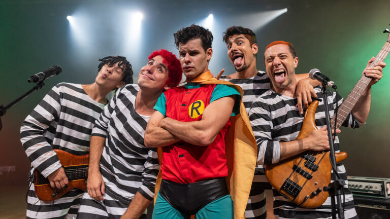 Atores de Mamonas Assassinas posam no palco com roupas de presidiário e Robin fazendo caretas em cena do especial da Record