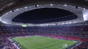 Vista interna do Estádio Ahmad bin Ali iluminado com gramado verde e arquibancadas vermelhas em noite de jogo de futebol no Catar
