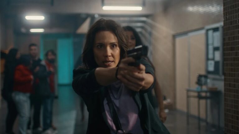 Mulher com expressão tensa segura arma apontando à frente em corredor com pessoas assustadas ao fundo